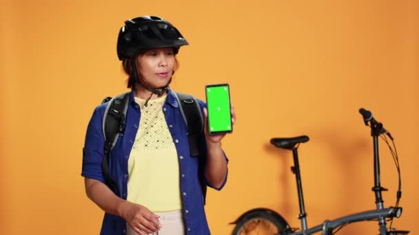 手持彩色键绿色屏幕智能手机的信使 显示大拇指向上的手势 亚洲女人头戴防护头盔 背负保暖食品背包 与橙色工作室背景隔离 — 图库视频影像