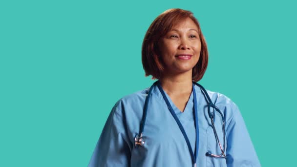 カメラを見ながら笑顔で笑顔を見せる熟練の美少女看護師 青のスタジオの背景に隔離されたプロのスクラブを身に着けている陽気なアジアの医療専門家の肖像 — ストック動画