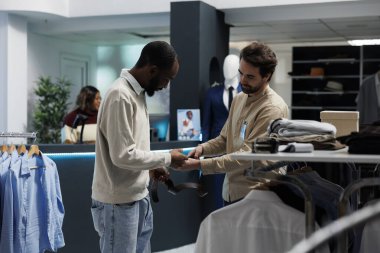 Giyim mağazası müşterisi ve işçi deri kemer kalitesini ve tarzını inceliyor. Afro-Amerikalı bir adam, erkek giyim mağazası alışverişi yaparken elinde aksesuar tutuyor ve butik asistanından tavsiye istiyor.