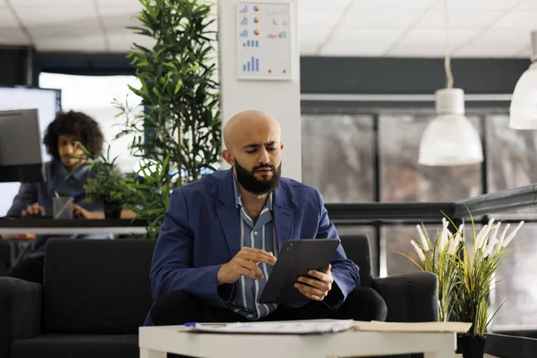 阿拉伯执行经理在办公室工作时检查平板电脑上的财务数据 创业公司员工在数字设备上浏览互联网 并坐在同事间的沙发上 — 图库照片