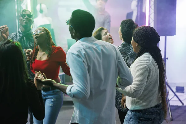 Diverse people clubbing and dancing together at discotheque party event in nightclub. Young friends holding hands, singing and relaxing on crowded dancefloor at social gathering
