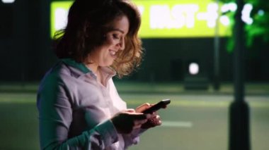 Gülümseyen kadın mesajlar atıyor, şehir merkezinde dolanıyor, mesajlar okuyor ve gece gezintisinden zevk alıyor. Sokak lambalarının altındaki gökdelenlerin yakınındaki akşam yürüyüşünde dinlenen biri. El kamerasıyla..