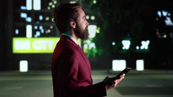 夜間の若者はスマートフォンを使って歩き 市内中心部を見回して高層ビルを鑑賞する ストリートライトの下を歩いている携帯アプリのビジネスマン ハンドヘルドショット — ストック動画