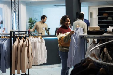 Kadın tişörtünü askıda tutuyor ve giyim mağazasında kıyafet seçerken kameraya bakıyor. Gülümseyen Afro-Amerikan müşteri butik portreden resmi kıyafet ve alışveriş seçiyor.