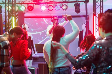 DJ, gece kulübünde sahnede çalarken mikrofonla dinleyicilere sesleniyor. İnsanlar dans ederken ve dans pistinde el kaldırırken kulüpteki elektronik müzik konserine katılıyorlar.