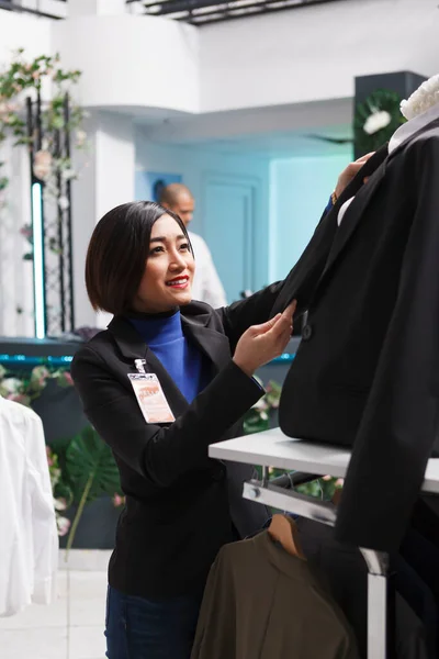 Retail center boutique asian woman employee adjusting jacket on mannequin while organizing womenswear for sale. Clothing store worker examining formal wear while managing inventory