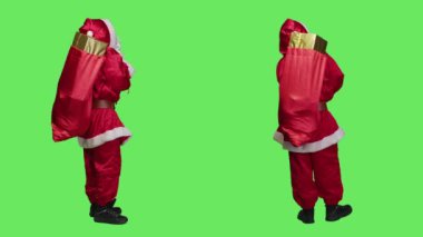 Noel Baba, stüdyoda, çocuklara oyuncak dağıtmaya hazırlanan yeşil perde arkaplanlı kırmızı hediye kutuları taşıyor. Bir çanta dolusu hediyeyle Aziz Nick 'i canlandıran genç bir yetişkin..