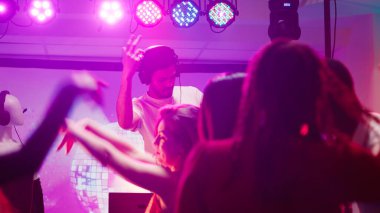 Erkek DJ kulüpte insanlarla parti yapıyor, diskotekte eğlenceli parti atmosferi yaratmak için ses istasyonunda müzik karıştırıyor. Genç adam dans ediyor ve arkadaşlarıyla zıplıyor, dans pistinde eğleniyor..