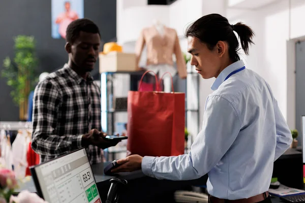 African american shopper paying for discount clothes with credit card at counter desk in clothing store. Shopaholic man shopping for casual wear, buying fashionable merchandise in modern boutique