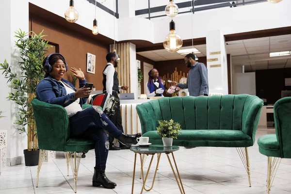 Happy woman traveler waving hand hello at smartphone camera, talking to family or friends on video call while traveling, sitting in hotel lobby. Internet communication during travel