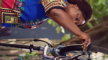 Hevesli siyahi bir kadının bisiklet tekerindeki hasarı dikkatle incelediği dikey video görüntüleri. Genç Afrikalı Amerikalı kadın bahçede bisikletini tamir ediyor, pedalları kontrol ediyor.
