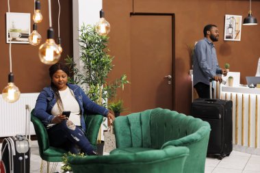 Otel lobisinde oturan Afrikalı Amerikalı kadın gezgin otel girişini beklerken arkadaşlarıyla internet üzerinden sohbet ediyor. Bavullu turistler resepsiyonda bekliyor.