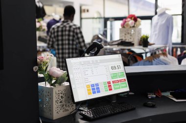 Kasası olan bilgisayarın seçici odak noktası modern butikte günlük kıyafet alışverişi yapan arka plandaki müşteriler. Afrikalı Amerikalı bir adam şık kıyafetlerle dolu askılara bakıyor.