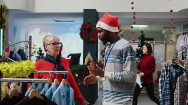 Çalışan, doğru tişörtü seçmiş yaşlı bir kadına Noel tatili için hediye olarak yardım ediyor. Müşteri kostüm mağazasında kıyafetleri karıştırıyor.