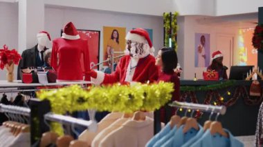 Çalışanlar Noel Baba kostümü giyerek bayram neşesini yaymak için alışveriş merkezindeki giyim mağazasına gidiyorlar. Şenlik temalı takım elbiseli işçi kış tatili sezonunda müşteriye kırmızı giysi gösteriyor.