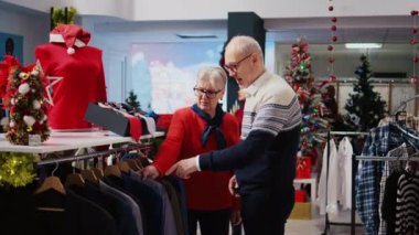 Yaşlı karı koca Noel alışverişi sırasında kostüm mağazasında elbise raflarına göz atıyorlar. Resmi kıyafetler almak isteyen yaşlı müşteriler, tatil promosyon satışlarının keyfini çıkarıyorlar.