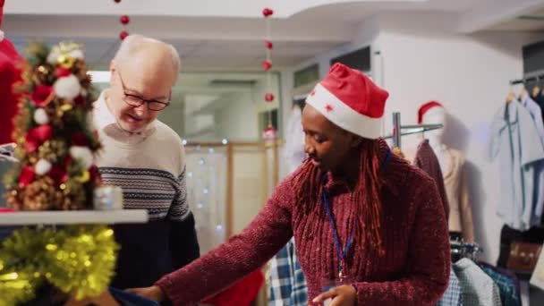 お祝いのホリデーシーズンにサンタの帽子をかぶっている便利な小売アシスタントは 最高のフィッティングブレーザーを持つ老人を助けます クリスマスの装飾された衣料品店で顧客を支援する従業員 — ストック動画
