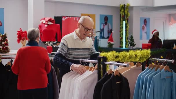 圣诞佳节期间 年事已高的顾客在节日华丽时尚精品店的衣架里浏览一番 在圣诞家庭活动中 这对老年夫妇在找到彩色服装准备送礼后 感到很快乐 — 图库视频影像