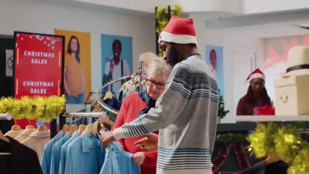 サンタの帽子をかぶっているアフリカ系アメリカ人労働者は お祝いのクリスマスの装飾された衣料品店で服を着る女性を助けます お祝いのファッションショップで高齢者のお客様を支援する従業員 — ストック動画