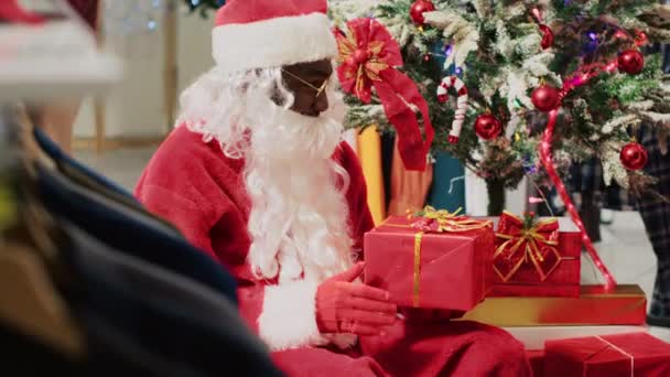 お祝いのプロモーションイベント中にクリスマスツリーの隣に座って サンタクロースの服を着た俳優と話すファッションストアの顧客 ホリデーシーズン中にワーカーからプレゼントを受け取るクライアント — ストック動画