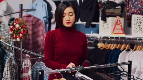 アジアの顧客は 服のサイズをチェックして クリスマステーマの店で服の棚を通ってブラウジングします 冬のホリデーシーズン中にXmasで女性がファッションブティックを飾る — ストック動画