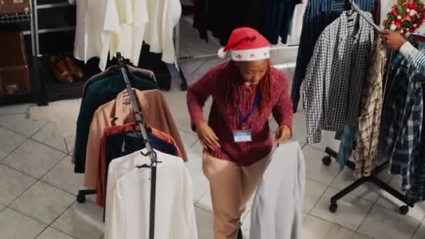 サンタの帽子をかぶっている従業員や お祝いの休日のシーズン中にクリスマスの装飾衣料品店を歩く顧客 Xmasのファッションショップの労働者とクライアント — ストック動画