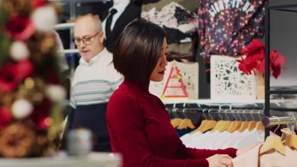 女人在圣诞装饰品商店的架子上浏览精美的夹克 寻找完美的圣诞晚宴服装 时尚的顾客需要新衣服 — 图库视频影像