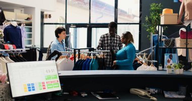 Store worker helping couple with trendy clothes, showing hangers with new fashion collection in modern boutique. African american customers shopping for stylish merchandise in clothing centre clipart