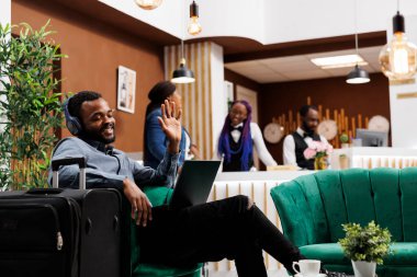 Mutlu gülümseyen Afrikalı Amerikalı girişimci otel lobisinde bavuluyla oturmuş kayıt beklerken bilgisayardan el sallıyor. Seyahat ederken konferans çağrısına katıl