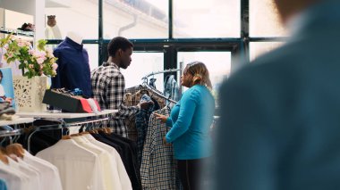 Afro-Amerikan çift şık ürünlere bakıyor, giyim mağazasındaki kumaşlara bakıyor. Gülümseyen müşteriler alışveriş yapmaktan hoşlanıyor, modern butikten moda kıyafetler ve aksesuarlar alıyorlar.
