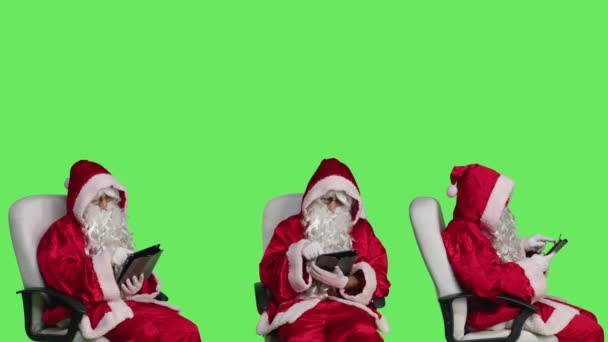 セントニックはグリーンスクリーン上のタブレットで動作し ウェブページをオンラインで閲覧し クリスマスイブの休日を祝う準備をしています 椅子に座っているデバイスを使用してサンタクロースの有名な衣装の若い大人 — ストック動画