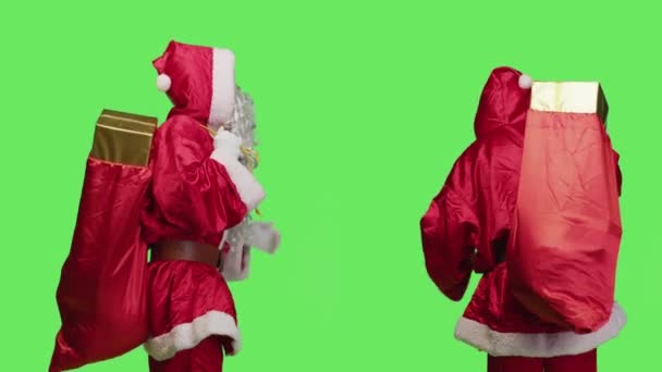 父のクリスマスはスタジオで象徴的なおもちゃ袋を運び 子供のためのリボン付きの箱にプレゼントを届けるために働いています 孤立した緑色の背景の上にポーズギフトの巨大な袋を持つサンタクロース — ストック動画
