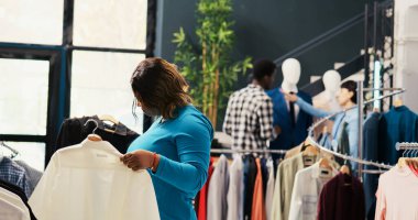 Yeni moda koleksiyonuna bakan Afrikalı Amerikalı müşteriler modern butikte şık kıyafetler alıyorlar. Moda ürünleri ile rafları kontrol eden şık bir istemci, mağazadaki kumaşı analiz ediyor