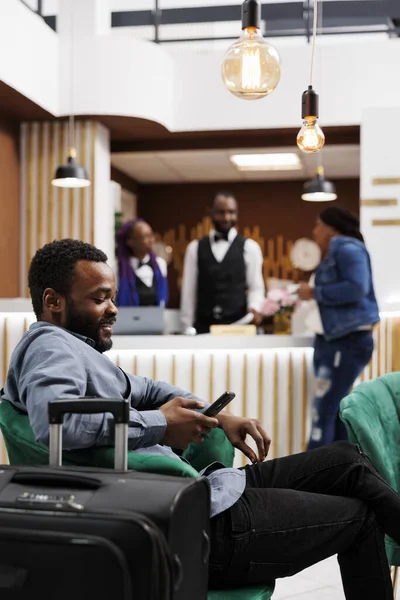 非洲裔美国人千禧年游客 带着行李坐在舒适的酒店大堂里 看着智能手机屏幕 在国际上使用手机 旅行时与朋友保持联系 四处漫游 — 图库照片