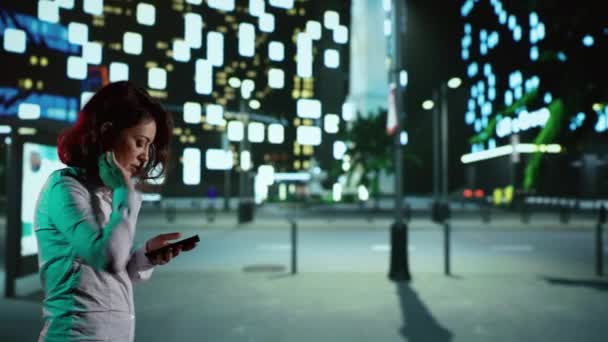 时尚人士用智能手机来寻找方向 带着路灯和高大的办公楼在市中心四处走走 穿着西服的女商人在夜间散步时感到快乐 — 图库视频影像