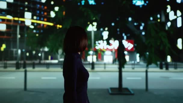 街の中心部近くを散策するアジア人で 街灯の下の歩道を歩いている間に近代的な高層ビルを見ています 夜のダウンタウンに一人でいる女性 遊歩道で微笑む — ストック動画