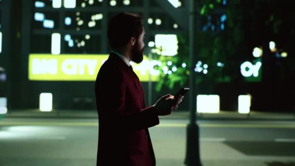 夜に街を歩き回るスタイリッシュな男が 街の高い建物の近くをライトで散策する 起業家は街灯で照らされた街の中心部の高層ビルの近くを歩き 電話でメッセージを送ります — ストック動画