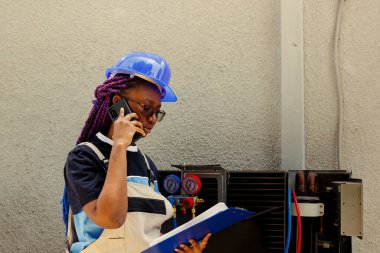 Havalandırma parçalarının değiştirilmesi gerektiği konusunda uzman teknisyen Afrikalı Amerikalı çırak telefonla bilgilendiriyor. Hava filtrelerini temizledikten sonra tamirci bakım planını kontrol ediyor.