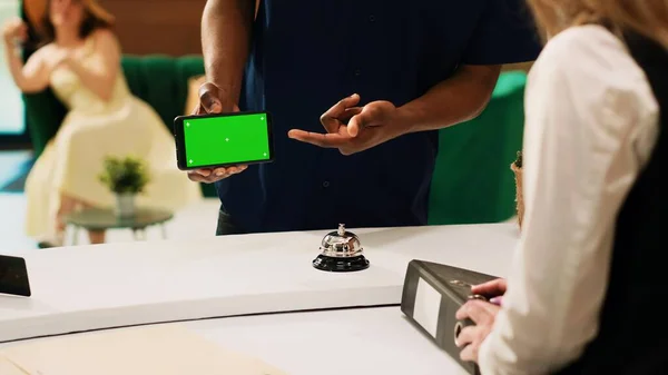 緑の画面で電話を示すゲストは ロビーのホテルのスタッフに隔離されたクロマキーディスプレイと携帯電話のテンプレートを保持します 男旅行者使用モックアップコピースペースでフロントデスク — ストック写真