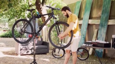 Sağlıklı erkek bisikletçi dikkatle bisikletin hasarlı lastiğini gevşetiyor ve söküyor. Sportif genç adam dikkatli bir şekilde bisiklet tekerleğini çözüyor ve ön çatalı ev bahçesine yerleştiriyor..