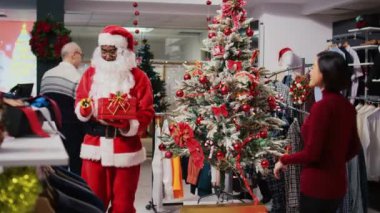 Afrika kökenli Amerikalı bir çalışan Noel Baba kostümü giyiyor. Güzel dekore edilmiş Noel ağacının yanında duruyor. Şenlik temalı kıyafet mağazasında, meraklı Asyalı müşteriyle konuşuyor.