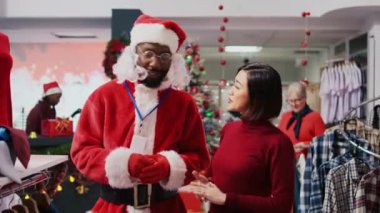 Noel Baba kostümü giyerek bayram neşesini yaymak için alışveriş merkezindeki giyim mağazasının etrafında perakende satış görevlisi. Xmas temalı takım elbiseli çalışan mutlu müşteri kırmızı giysisini gösteriyor