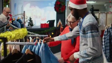 İşçi, gelecek Noel tatilinde kocasına hediye olarak vermek üzere doğru kıyafeti seçen kıdemli müşteriye yardımcı oluyor. Şenlikli moda dükkanında müşteriler kıyafetleri karıştırıyor.