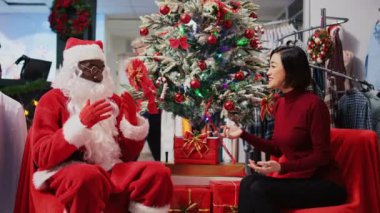 Noel Baba gibi davranan işçinin Noel ağacının yanında Xmas dekore edilmiş giyim mağazasında Asyalı bir kadınla oturması. Moda mağazası perakende asistanı müşteriye hediye sunuyor