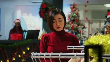 Mutlu Asyalı kadın portresi, Noel tatilinde alışveriş merkezinin kıyafet mağazasında elbise raflarında geziniyor. Moda mağazasından hediye almak isteyen bir müşteri.