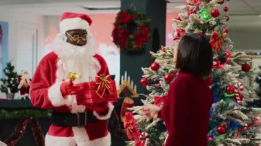 Noel Baba gibi giyinmiş Afrikalı bir çalışan Noel Baba zili çalıyor ve elinde Xmas süslü ağacının yanında Xmas hediyesiyle Asyalı müşteriyle şenlikli moda mağazasından alışveriş yapıyor.