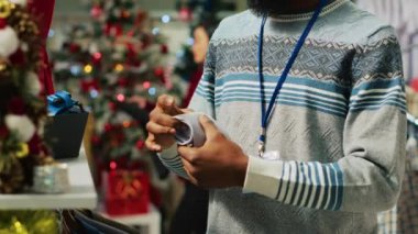 Gülümseyen bir işçinin portresi Noel süslü kıyafet mağazasında kravat düzenlerken. Neşeli moda işçisi tatil sezonu alışverişi için mal hazırlıyor.
