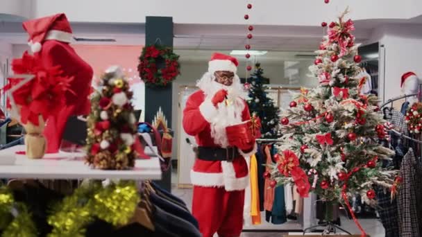 アフリカ系アメリカ人男性は クリスマスにサンタクロースをテーマにしたショッピングモールの衣料品店で服を着ていました 従業員のジングリングXmasの鐘およびお祝いの装飾された店に存在する赤いパッケージを握って下さい — ストック動画