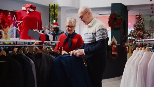 クリスマスショッピングのスリーの間にお祝いの服の店で服ラックをブラウジングシニアカップル 正式な服装を購入する予定の古いクライアント 休日プロモーション販売を楽しむ — ストック動画