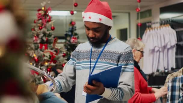 アフリカ系アメリカ人の従業員は クリスマスの装飾されたファッションストアで衣服ラックを閲覧し クリップボードの調整価格を書き留めています 労働者がブレーザーを検査し 損害賠償を求める — ストック動画